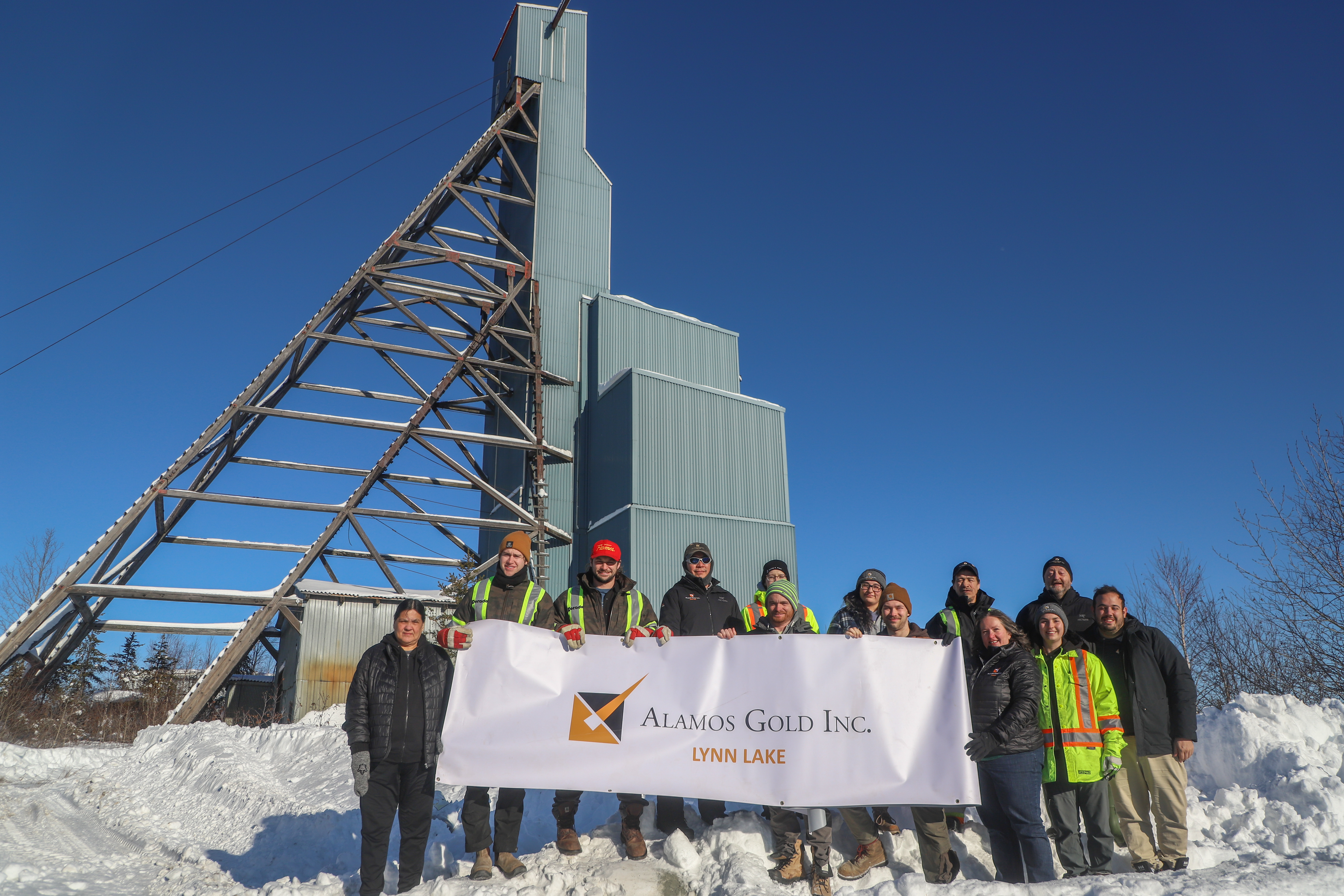 Group of people standing next to each other in the snow in front of a building and holding an Alamos Gold Lynn Lake banner.