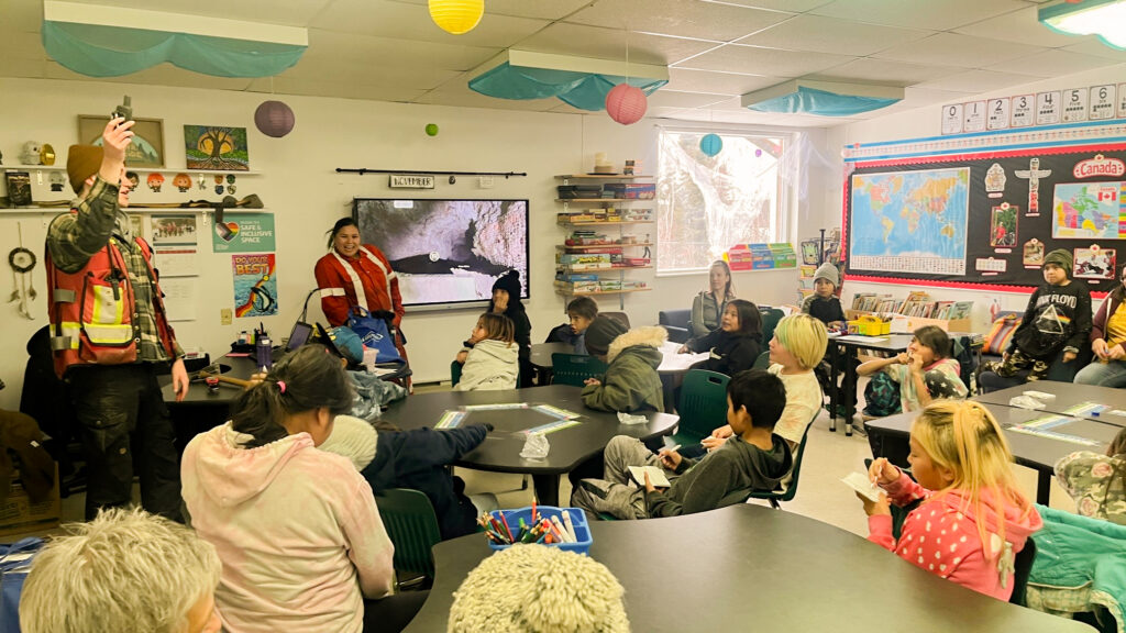 Image of a classroom with several students siting on their desks and 2 people are at the front speaking, one of them is holding something in one hand.
