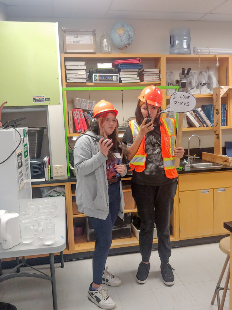 2 students wearing hard hats and holding other tools and equipment, one of them is holding a sign that says 'i love rocks'.